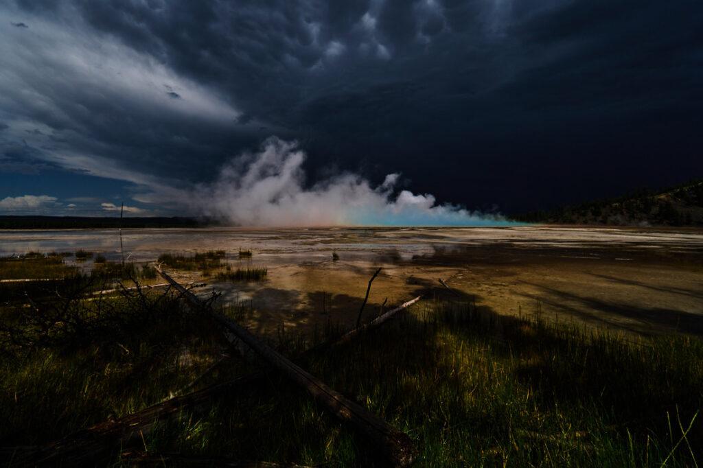 Grand Prismatic Spring in Yellowstone National Park viewed from the side after a passing storm, its famous colors visible in the steam, crossed logs in foreground